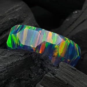 Pode incluir: Um anel facetado com uma base escura e flashes iridescentes vibrantes de verde néon, laranja e azul. A superfície da banda é composta por múltiplos planos geométricos planos que captam a luz, criando um jogo de cores sobre o material escuro.