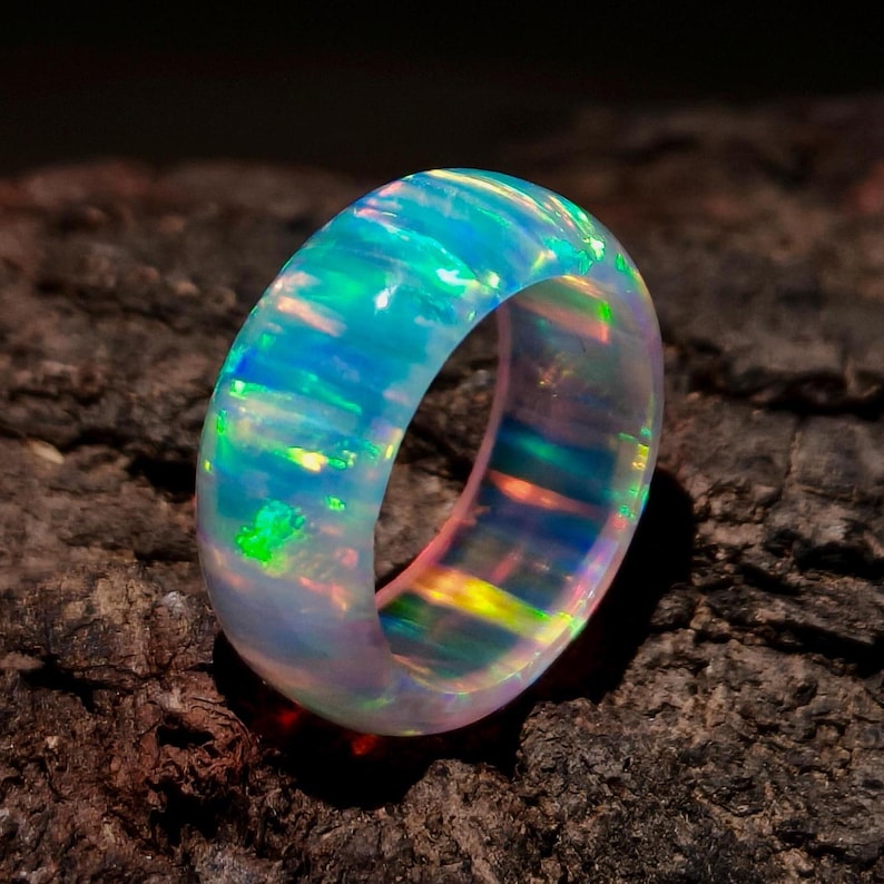 Puede incluir: Un anillo de &oacute;palo luminoso con una vibrante exhibici&oacute;n de colores, incluyendo azules, verdes y toques de amarillo y naranja. La superficie iridiscente del anillo captura la luz, creando una pieza de joyer&iacute;a cautivadora y &uacute;nica.