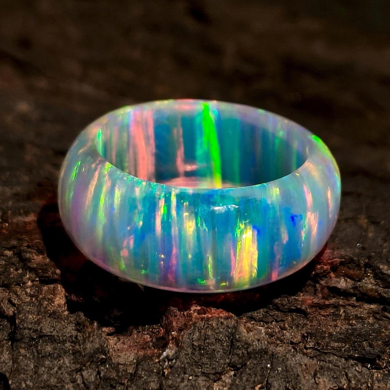 Puede incluir: Un anillo ancho de &oacute;palo iridiscente. El anillo muestra un espectro de colores, incluyendo azules, verdes y rosas, con una superficie pulida y lisa. El &oacute;palo se encuentra sobre un fondo oscuro y texturizado.