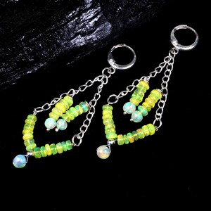 Puede incluir: Un par de pendientes colgantes con un diseño de cadena plateada. Los pendientes están adornados con pequeñas cuentas de color verde claro y amarillo, y una sola cuenta iridiscente opalina más grande en la parte inferior. El fondo es negro.