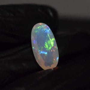 Puede incluir: Una piedra preciosa de ópalo ovalada blanca con destellos iridiscentes verdes, azules y naranjas.