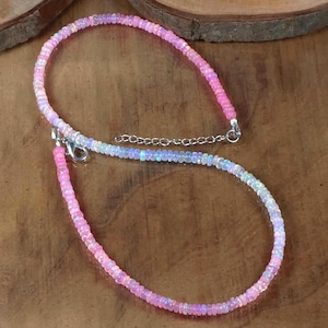 May include: A delicate necklace featuring small, iridescent opal beads in shades of pink, white, and blue. The necklace has a silver clasp and chain, and is displayed on a wooden surface.