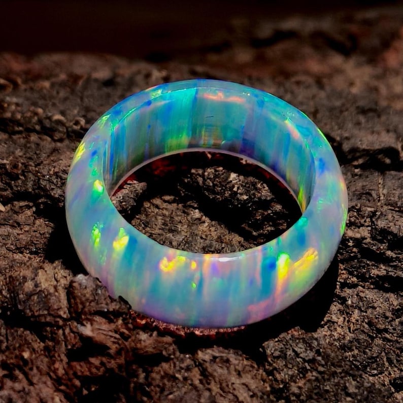 Puede incluir: Un vibrante anillo de &oacute;palo, que muestra un espectro de colores que incluye azules, verdes y amarillos. El material transl&uacute;cido del anillo permite el paso de la luz, creando un efecto luminoso. El anillo est&aacute; sobre un fondo oscuro y texturizado.