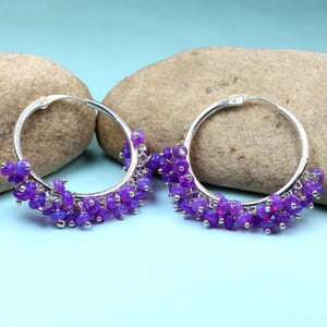 May include: Silver hoop earrings with a fringe of small, purple beads. The earrings are displayed against a light blue backdrop, with two large, light brown rocks in the background.