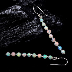 Op de afbeelding: Een paar oorhangers met iriserende opaal kralen geregen op zilverkleurige haken. De opalen tonen een reeks kleuren, waaronder roze, blauw en groen. De oorbellen zijn ontworpen om naar beneden te hangen. De achtergrond is donker.