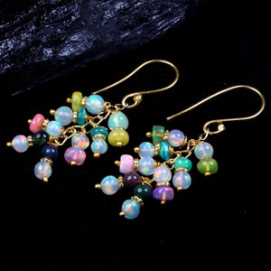 May include: A pair of gold-colored dangle earrings. Each earring features a cluster of colorful beads, including opal, green, pink, and blue, suspended from a curved hook. The beads vary in size and shape, creating a playful and eye-catching design.