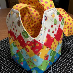 Puede incluir: Una bolsa de mano de patchwork colorida con un patrón amarillo, blanco, rojo, azul y verde. La bolsa tiene un fondo blanco con varias imágenes de arcoíris, soles, fresas y ranas verdes. La bolsa tiene dos asas amarillas.