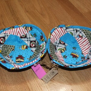 May include: Two blue fabric bowls with a patchwork design featuring red and white stripes, green honeycomb, and a blue background with white polka dots and cherries. The bowls are turned upside down and have a tag attached.