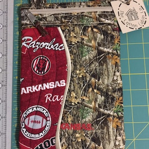 Op de afbeelding: Gewatteerde crossbodytas met een camouflagepatroon en een rood paneel met de tekst "Razorbacks" en "Arkansas" en logo's. De tas heeft een ritssluiting, een klein label met de tekst "The Cotton Cabin Co." en een metalen clip.