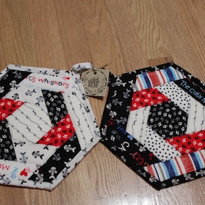 Pode incluir: Dois pegadores de panela hexagonais com um design de patchwork. Um pegador de panela é preto, branco, vermelho e azul com o texto "The Cat's Meow". O outro pegador de panela é preto, branco, vermelho e azul com o texto "It's Whiskers".