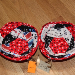 May include: Two red fabric bowls with a patchwork design featuring paw prints, fish bones, and the text "The Whisker" and "The Cat" in black and white.