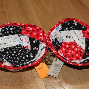 May include: Two red, black and white fabric bowls with a patchwork design. The bowls are made of fabric scraps with different patterns, including paw prints, fish bones, and the words "meow" and "the cats".
