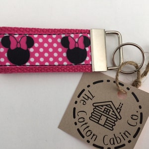 May include: Pink and white polka dot fabric key chain with two black and white Minnie Mouse heads. The key chain has a silver metal ring and a silver metal clasp. The key chain has a brown tag with the text "The Cotton Cabin Co."