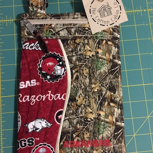 Op de afbeelding: Een rode en witte stoffen tas met een camouflagepatroon aan de achterkant. De tas heeft een ritssluiting en een band. De voorkant van de tas heeft de woorden "Razorback" en "Arkansas" samen met een varken en een voetbalhelm-afbeelding. De tas heeft een label met de tekst "The Cotton Cabin Co."