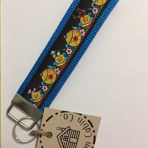 Pode incluir: Um chaveiro azul e preto com um design floral e um chaveiro de prata. O chaveiro tem uma etiqueta que diz "The Cotton Cabin Co."