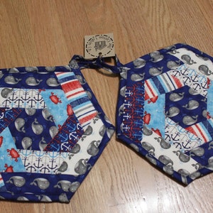 May include: Two blue and white quilted pot holders with a whale and anchor design. The pot holders are hexagonal in shape and have a red, white, and blue striped border.