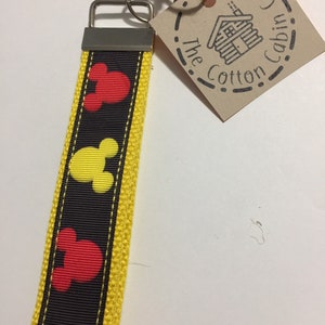 Puede incluir: Un llavero amarillo y negro con un patrón repetido de cabezas de Mickey Mouse rojas y amarillas. El llavero tiene un llavero plateado y una etiqueta marrón que dice "The Cotton Cabin Co."