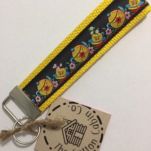 Pode incluir: Chaveiro amarelo e preto com uma fita preta e branca com um padrão repetido de bules e flores. O chaveiro tem um anel de chave prateado e uma etiqueta marrom com o texto "The Cotton Cabin Co."