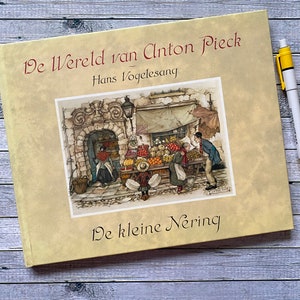 May include: A yellow book with the title "De Wereld van Anton Pieck" by Hans Vogelesang. The cover features an illustration of a street scene with a fruit stand and people.