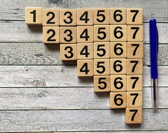 May include: Wooden number tiles arranged in a descending staircase pattern, displaying numbers 1 to 7. A blue pen rests vertically beside the tiles on a weathered, grey wooden surface. The numbers are printed in black.