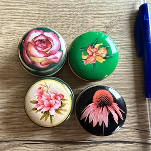 Può includere: Quattro contenitori rotondi decorativi con disegni floreali. I contenitori presentano immagini di una rosa, un fiore arancione, fiori rosa e una coneflower rosa. Una penna blu è accanto ai contenitori.