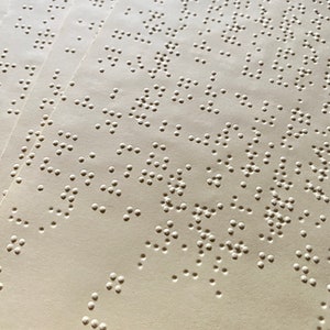May include: Close-up of a stack of beige paper with raised dots in a braille pattern.