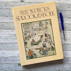 May include: A vintage book titled "ANTON PIECK'S SPROOKJESBOEK" with an illustration of a town scene. The book has a cream-colored cover and is accompanied by a blue pen. The book is resting on a light gray wooden surface.