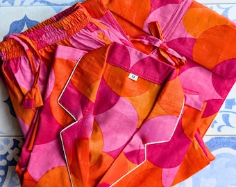 Pode incluir: Um conjunto de pijama rosa e laranja com um padrão geométrico de círculos sobrepostos. O conjunto inclui uma parte superior de manga comprida com colarinho e uma calça com cós elástico. A parte superior tem uma bainha branca ao redor do colarinho e dos punhos. O rótulo de tamanho na parte superior diz "M".