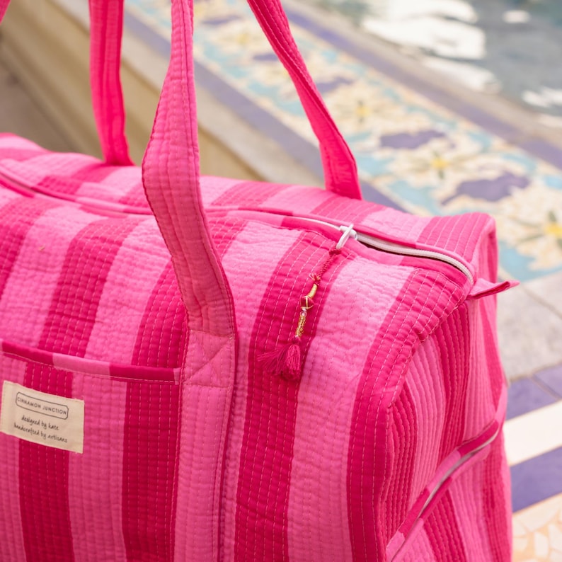 Pode incluir: Uma mala de viagem listrada rosa e f&uacute;csia com al&ccedil;as combinando e fecho de z&iacute;per. A mala tem uma pequena etiqueta retangular com texto. A mala &eacute; feita de tecido acolchoado.