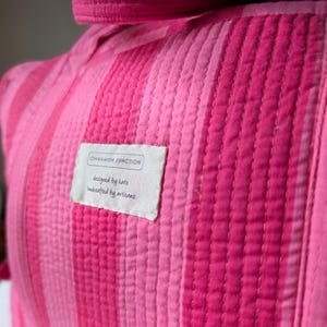 Pode incluir: Close-up de um tecido acolchoado com listras verticais em tons variados de rosa. Uma etiqueta retangular com o texto "CINNAMON JUNCTION" e "designed by kate, handcrafted by artisans" &eacute; costurada no tecido.