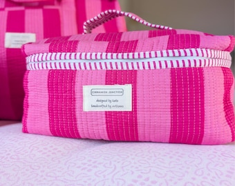 Pode incluir: Uma bolsa de cosméticos acolchoada com listras rosa e fúcsia com zíper listrado branco e azul. A bolsa tem uma alça e uma etiqueta que diz "Cinnamon Junction, designed by Kate, handcrafted by artisans."