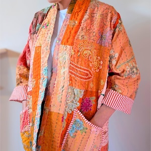May include: A patchwork kimono in shades of orange, peach, and pink, featuring floral and paisley patterns. The kimono has pockets and red and white striped cuffs. The garment is made of stitched fabric.