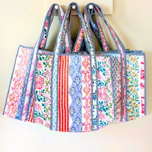 May include: Three colorful tote bags with floral and striped patterns. The bags have a rectangular shape with a striped handle and are hanging on a white wall. The bags feature a mix of pink, red, blue, and white fabrics.