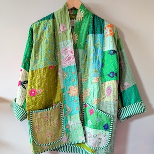 Op de afbeelding: Een patchwork kimono-achtige jas in groen- en tinten, versierd met geborduurde bloemen- en geometrische patronen. De jas heeft twee voorzakken en gestreepte randen op de manchetten en randen. Het hangt aan een houten hanger.