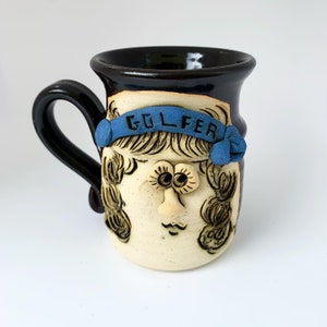 May include: A brown ceramic mug with a cartoon face and the word "GOLFER" on a blue headband. The mug has a brown handle and a white face with blue eyes and a brown hair.