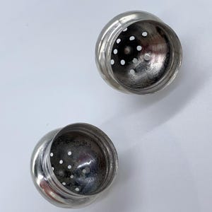 May include: Two silver metal salt and pepper shakers with a round shape and a perforated top. The shakers are empty and ready to be filled.