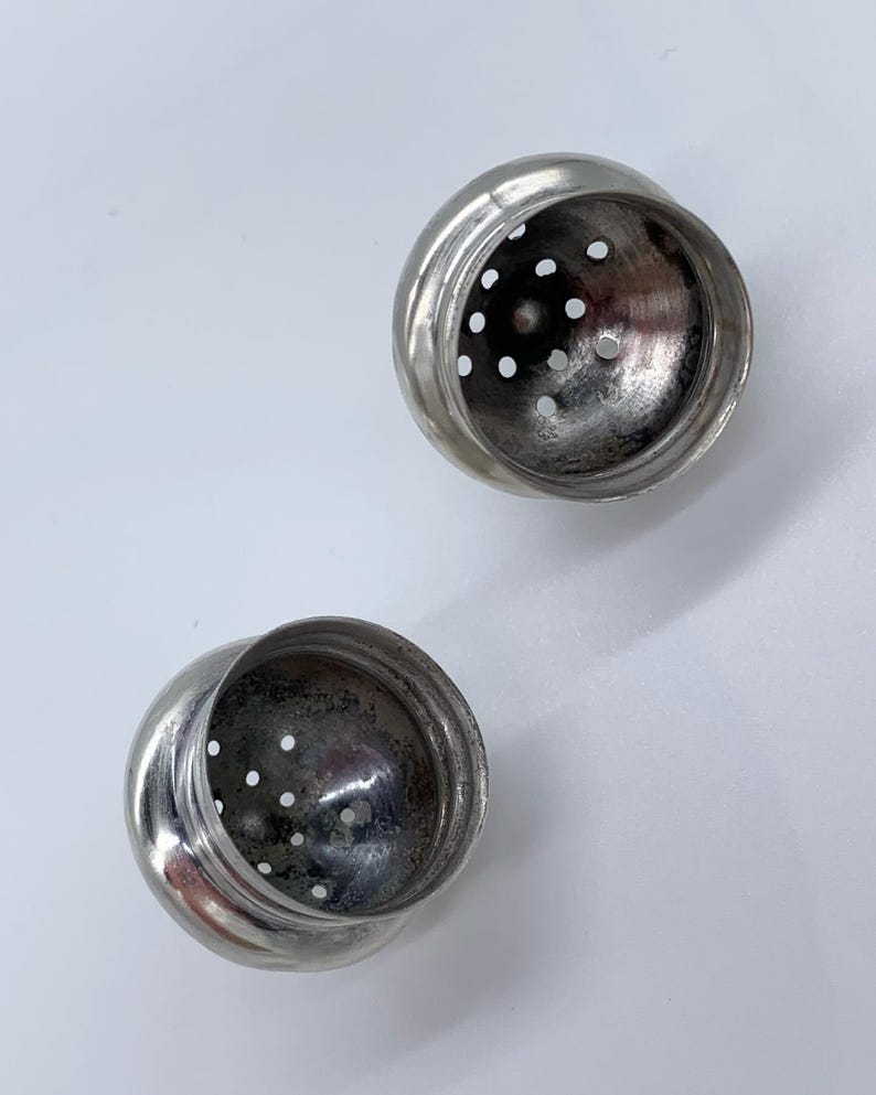 May include: Two silver metal salt and pepper shakers with a round shape and a perforated top. The shakers are empty and ready to be filled.
