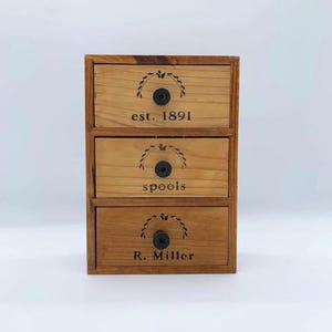 May include: A three-drawer wooden storage box with black knobs. Each drawer is labeled with text: "est. 1891", "spools", and "R. Miller". The box has a light brown finish and a decorative leaf design above each drawer.