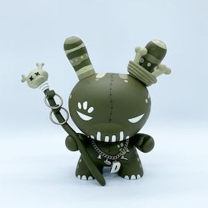 May include: A green and white toy rabbit with a skull and crossbones on its head, a crown, and a staff. The toy is wearing a necklace with a letter D pendant.