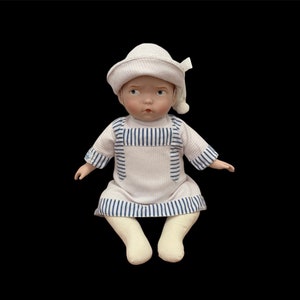 May include: A porcelain doll wearing a white and blue striped dress and a white hat. The doll has a painted face with blue eyes and rosy cheeks.