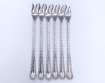 May include: Six silver-colored appetizer forks arranged vertically on a white surface. Each fork has a decorative handle and a four-pronged head. The forks are evenly spaced and appear to be made of metal.