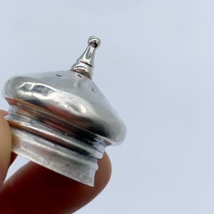 May include: Silver-toned metal bottle stopper with a pointed top and a round base. The stopper has a textured surface and several small holes.