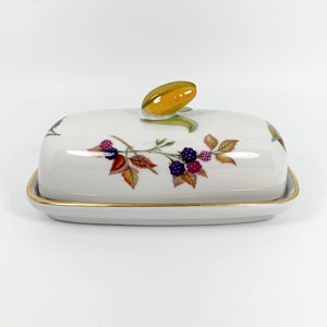 May include: A white porcelain butter dish with a gold trim and a floral design featuring blackberries and leaves. The lid has a yellow knob.