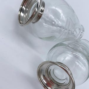 May include: Two clear glass bottles with etched designs and silver metal bases. The bottles are empty and have a vintage look.