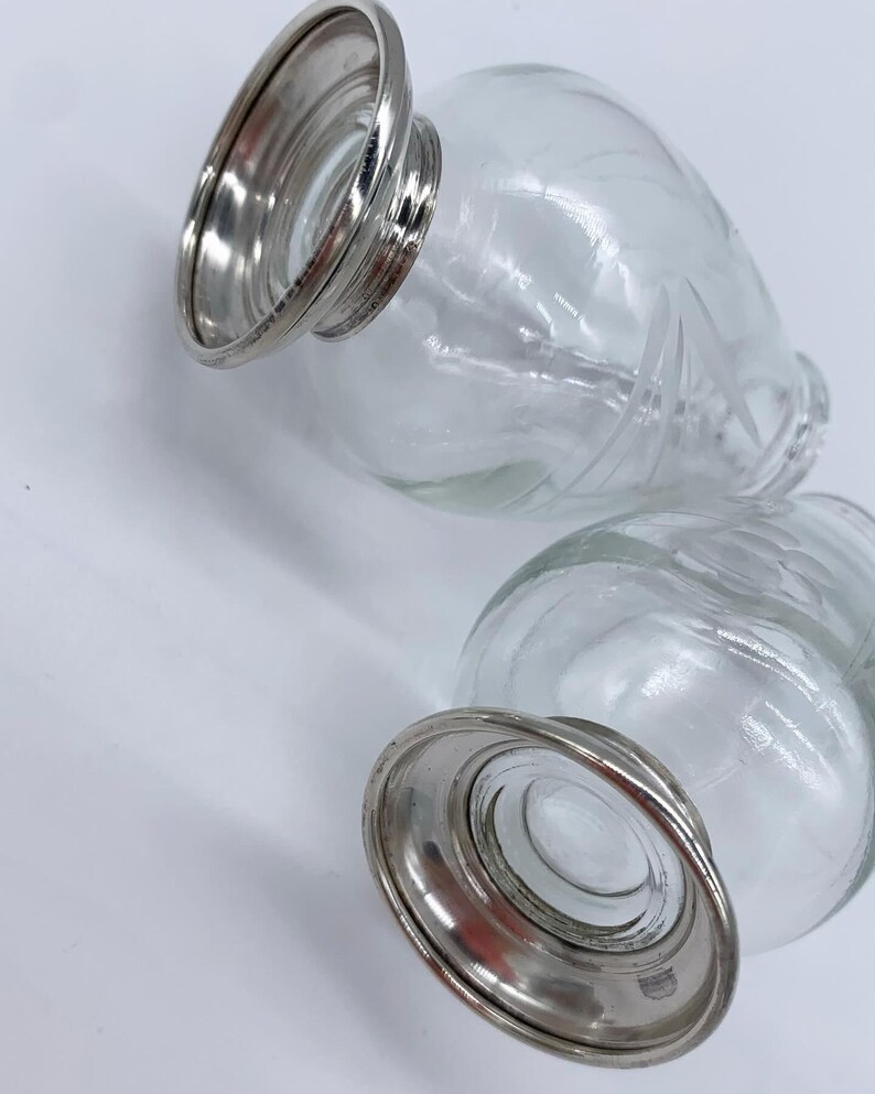 May include: Two clear glass bottles with etched designs and silver metal bases. The bottles are empty and have a vintage look.
