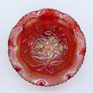 May include: A round, iridescent red glass bowl with a ruffled edge. The bowl features a raised floral design of roses and leaves in the center. The glass has a textured, shimmering finish, with hints of gold and purple.
