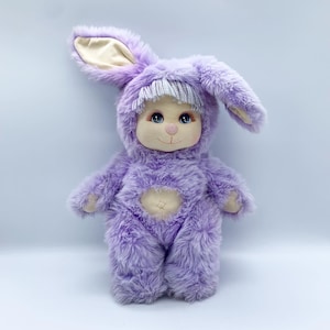 May include: A plush lavender doll in a bunny costume. The doll has a round face with blue eyes, a pink nose, and a small smile. The bunny costume features long ears, a hood, and a fluffy body. The doll's hands and feet are a lighter shade of lavender.