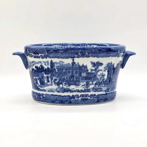 Peut inclure: Un bol en céramique blanche avec deux anses latérales, orné d'un motif bleu détaillé représentant un paysage avec des bâtiments, des arbres et des personnages. Le bord et la base présentent un motif géométrique et floral répétitif.