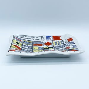 May include: White ceramic rectangular dish with a wavy edge. The dish is decorated with a colorful illustration of nautical flags and the text "FLAGS TO BE USED IN THE INTERNATIONAL CODE".