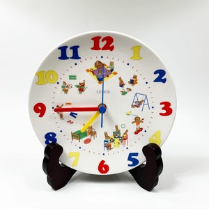 May include: A white ceramic clock with colorful numbers and a red and yellow hand. The clock features a design of children playing and the text "LENOX".